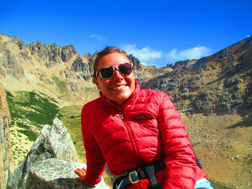 Emily in a red jacket and climbing harness, smiling on a granite ledge high above an alpine valley
